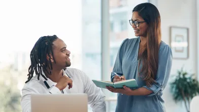 An optimistic looking man and woman collaborating at the office with a laptop and notebook.
