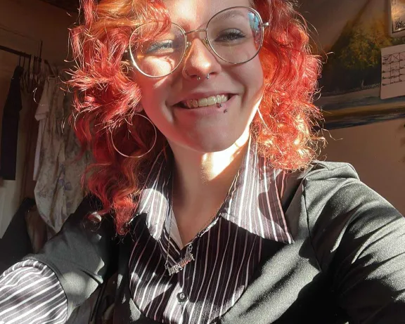 A selfie of a person with curly red hair and glasses smiling into the camera.